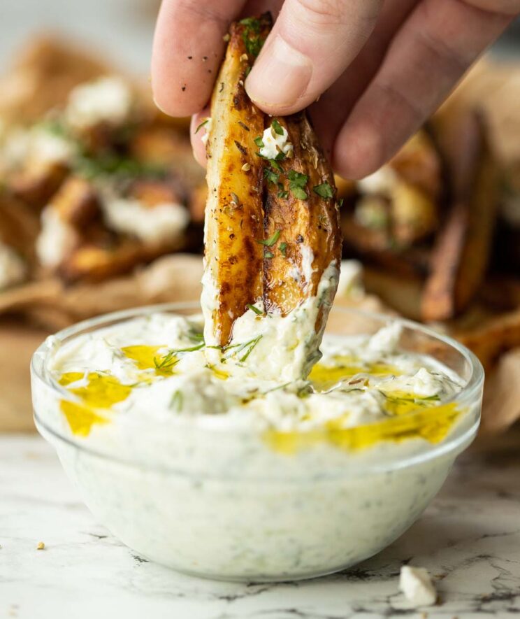hand dunking Greek fries into small glass bowl of Tzatziki