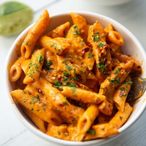 close up shot of pasta in small white bowl garnished with chilli flakes and parsley with lime blurred in background