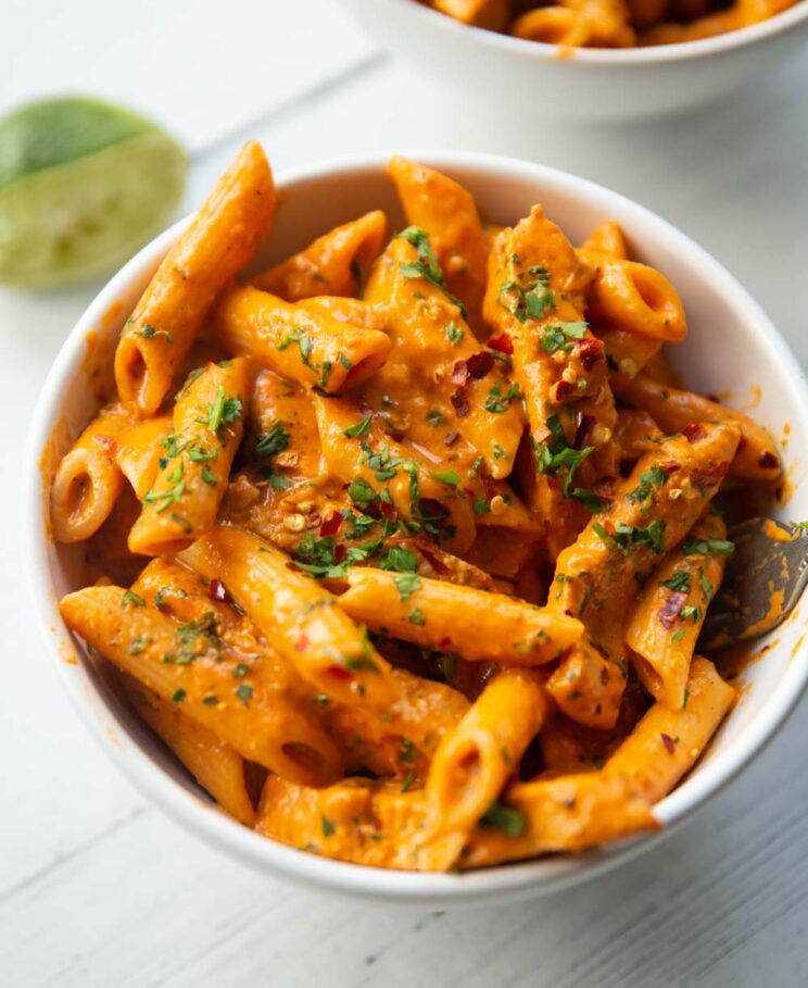 close up shot of pasta in small white bowl garnished with chilli flakes and parsley with lime blurred in background