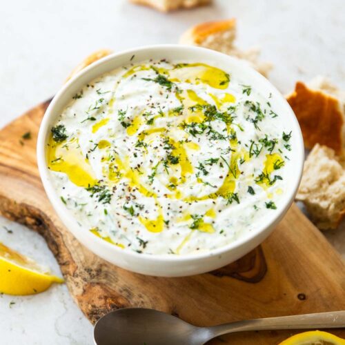 Tzatziki served in small white bowl on wooden chopping board surrounded by bread and lemon