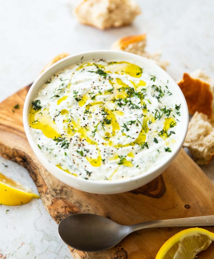 Tzatziki served in small white bowl on wooden chopping board surrounded by bread and lemon