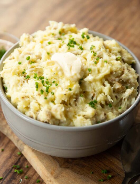 mashed potatoes in large grey bowl on wooden chooping board garnished with chives