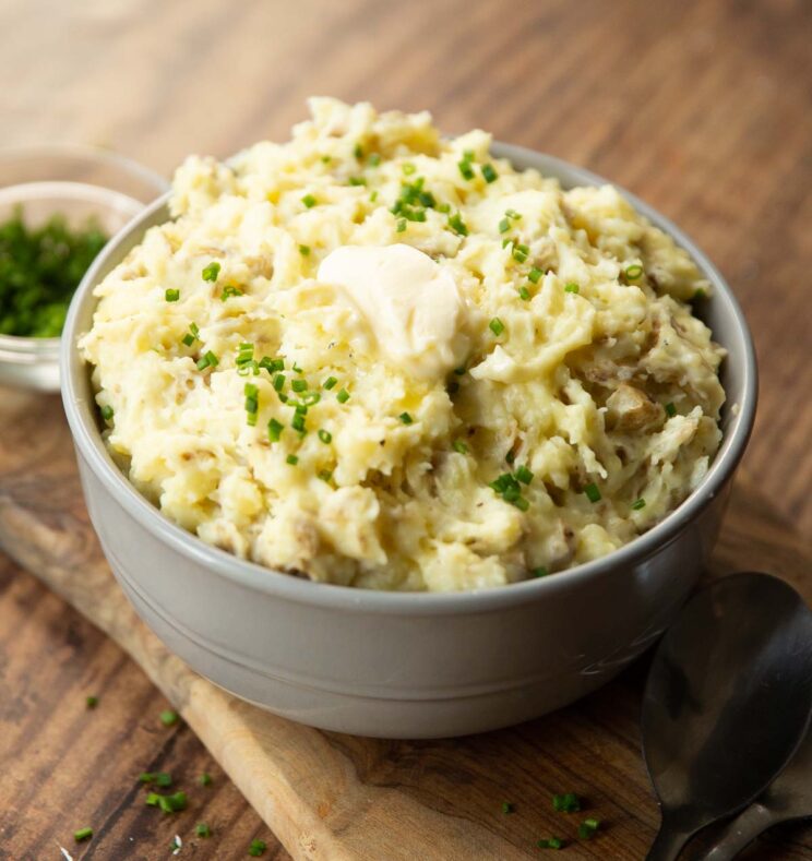mashed potatoes in large grey bowl on wooden chooping board garnished with chives