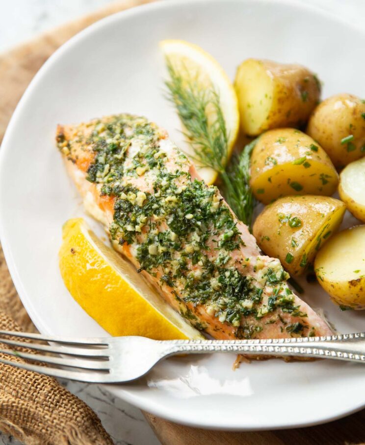 garlic herb salmon served on small white plate with baby potatoes, lemon wedges and silver fork