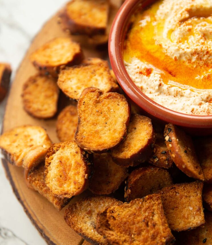 close up shot of bagel chips on wooden board served with bowl of hummus