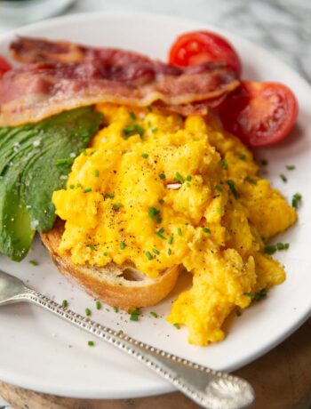 cheesy scrambled eggs served on toast on white plate with bacon, tomatoes and avocado