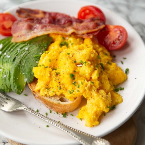 cheesy scrambled eggs served on toast on white plate with bacon, tomatoes and avocado