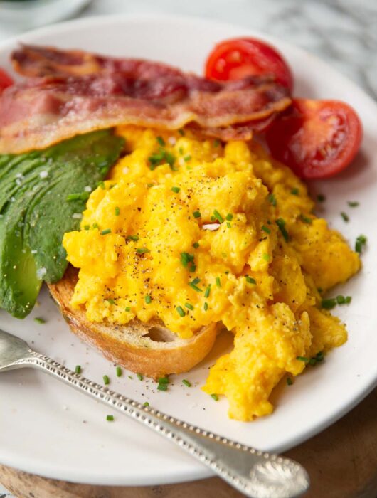 cheesy scrambled eggs served on toast on white plate with bacon, tomatoes and avocado