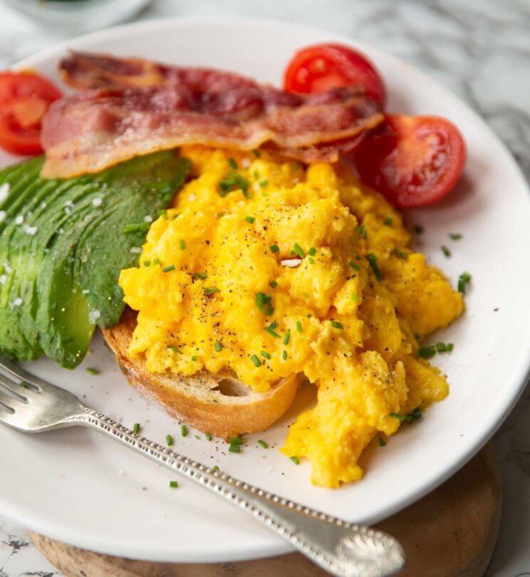 cheesy scrambled eggs served on toast on white plate with bacon, tomatoes and avocado