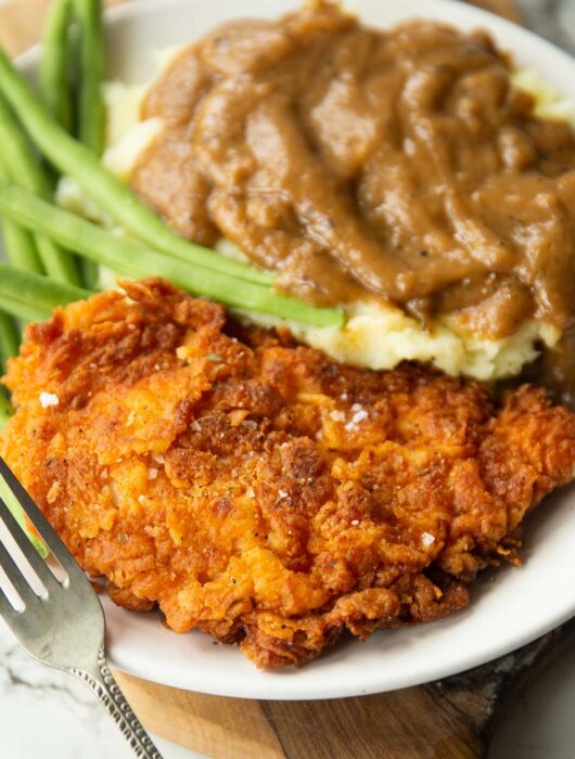 fried chicken cutlet served on small white plate with green beans, mash and gravy