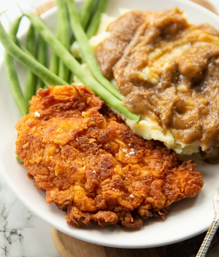 fried chicken cutlet on small white plate with mash, gravy and green beans blurred in the background