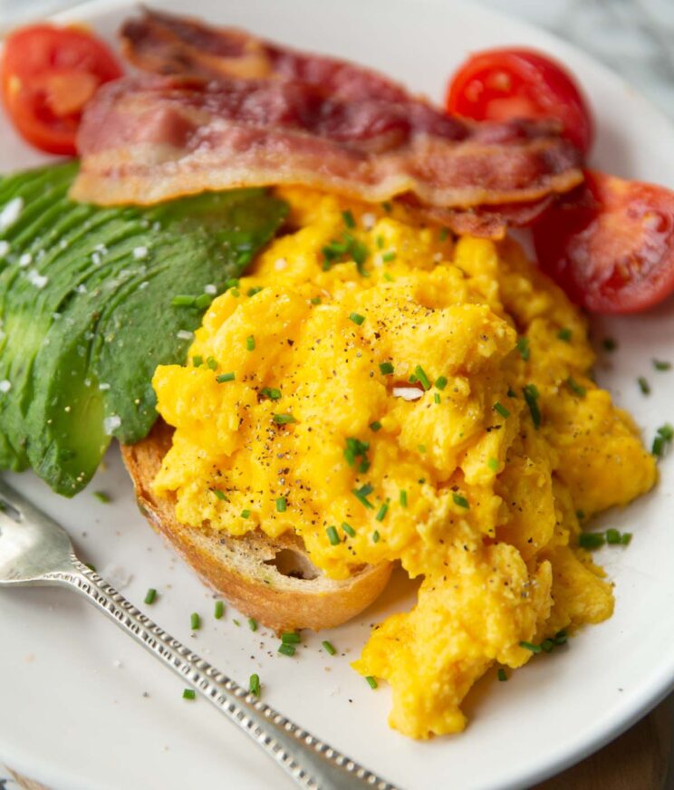 scrambled eggs with cheese on toast with bacon, avocado and tomatoes on white plate