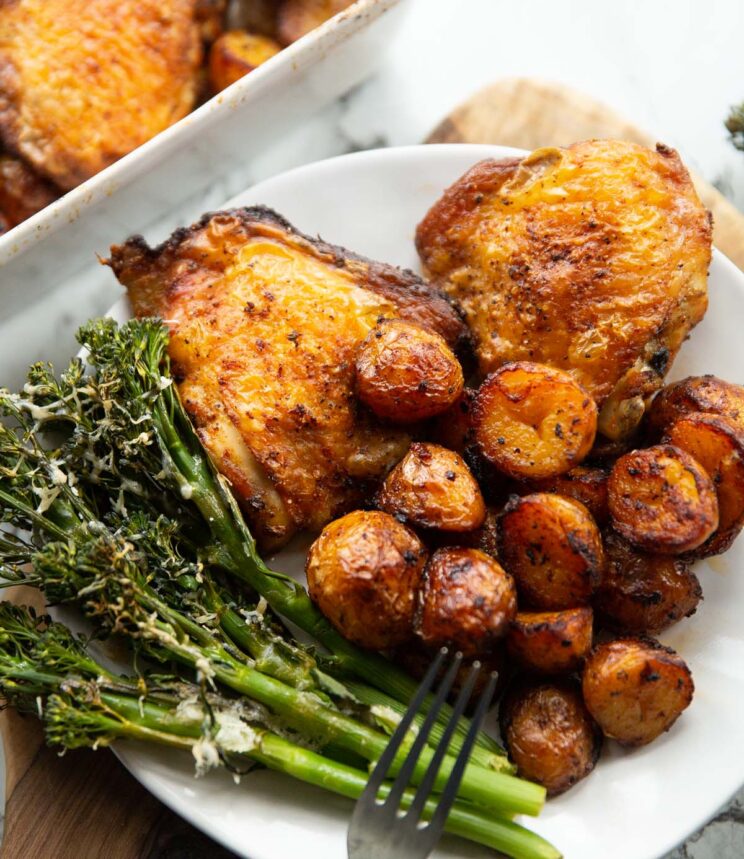 close up shot of baked chicken and potatoes on small white plate with broccolini