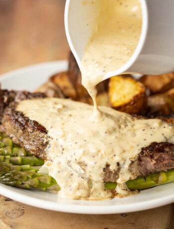 close up shot of small white pot pouring sauce over steak with potatoes and asparagus