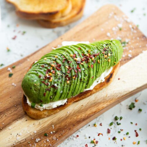 close up shot of avocado cream cheese toast on chopping board surrounded by garnish