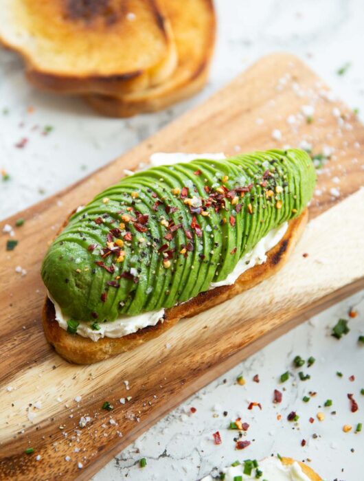 close up shot of avocado cream cheese toast on chopping board surrounded by garnish