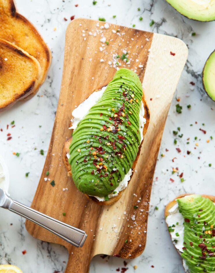 overhead shot of cream cheese avocado toast on chopping board surrounded by garnish