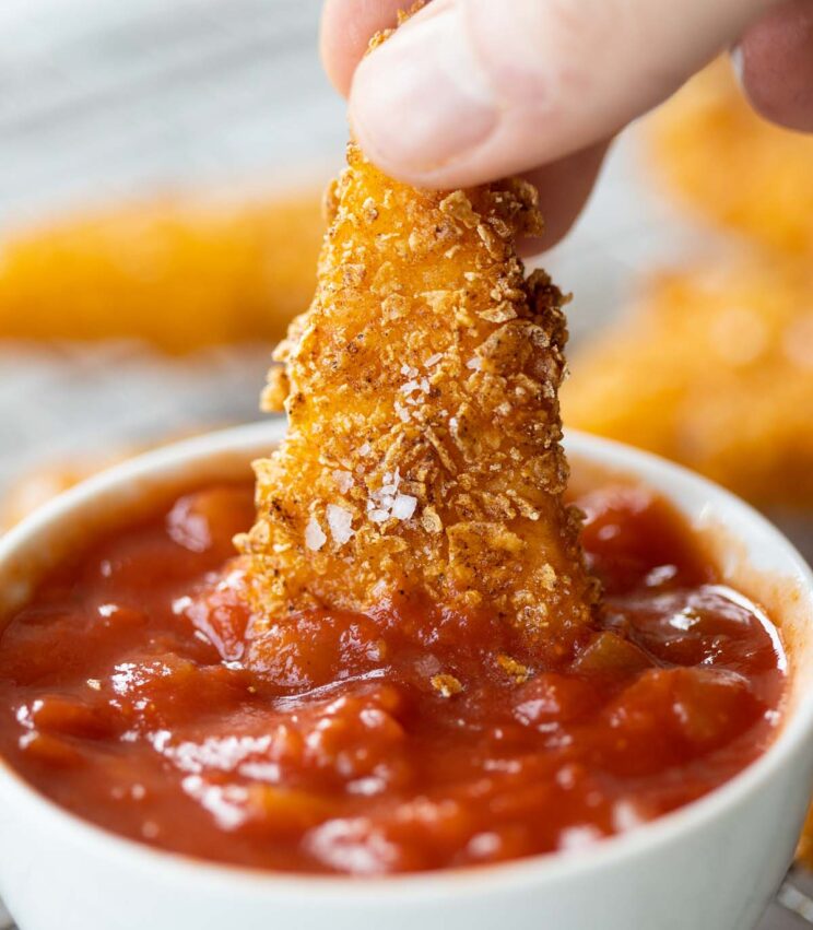 close up shot of fingers dunking Dorito crusted mozzarella into small white pot of tomato salsa