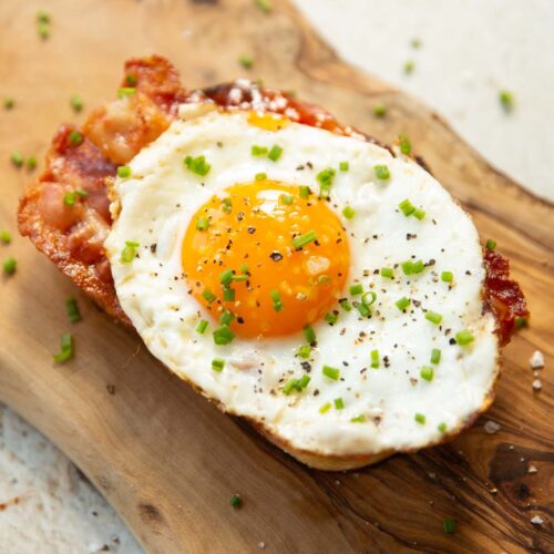 close up shot of egg and bacon on toast on wooden board garnished with chives