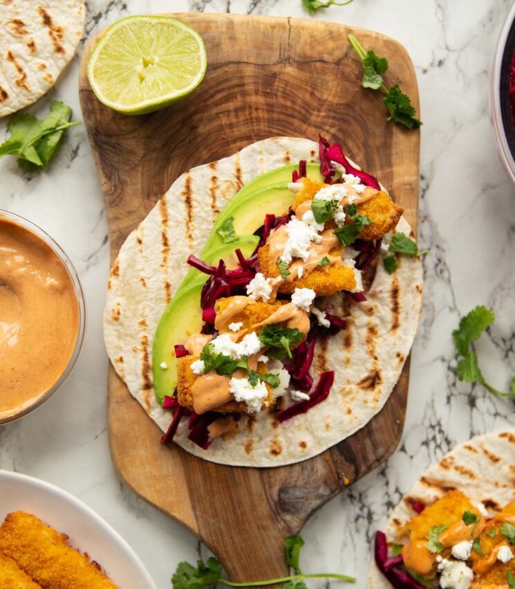 overhead shot of fish finger taco on wooden board surrounded by garnish
