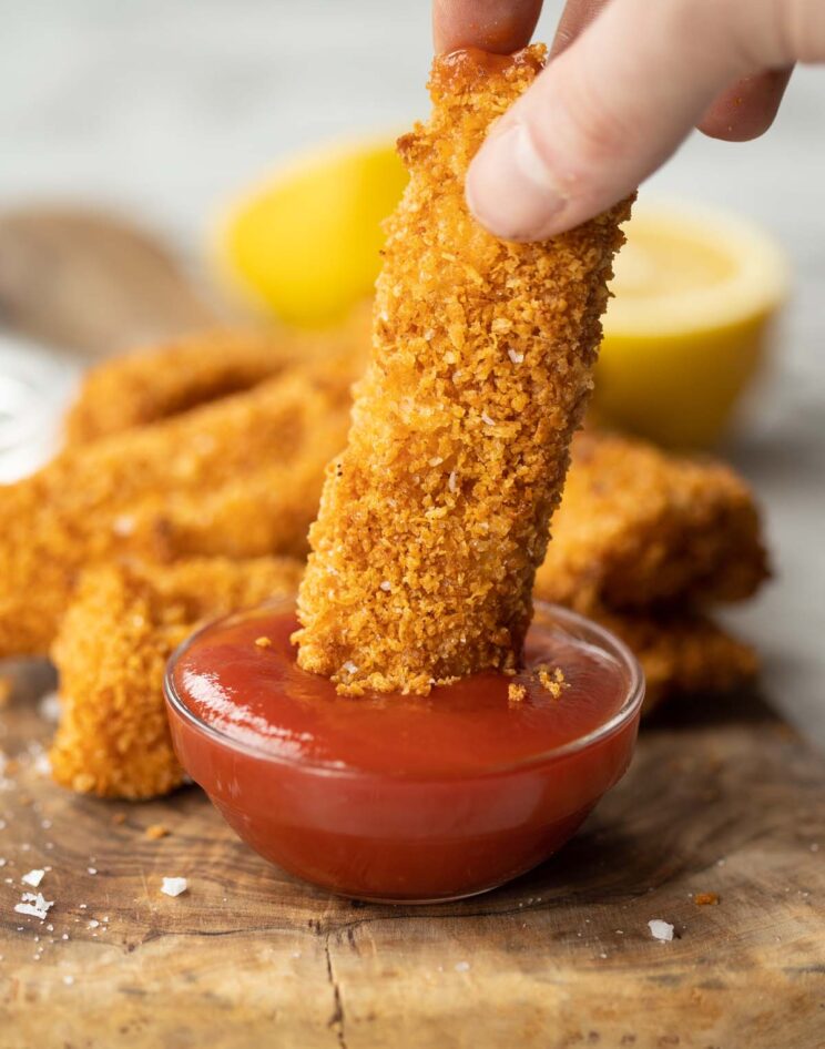 close up shot of hand dunking fish finger into small glass pot of ketchup