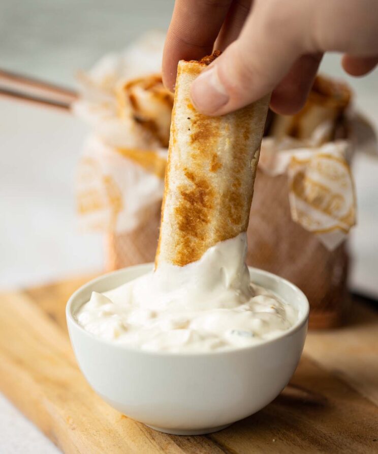 hand dunking a pizza roll up into small white pot of dip on wooden board