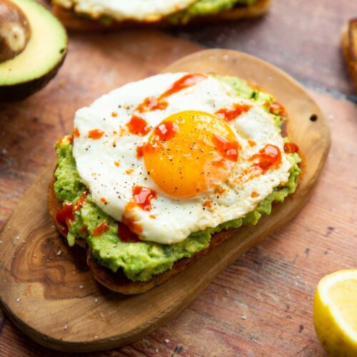 close up shot of avocado egg toast on small chopping board surrounded by garnish