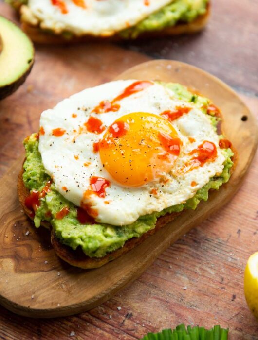 close up shot of avocado egg toast on small chopping board surrounded by garnish