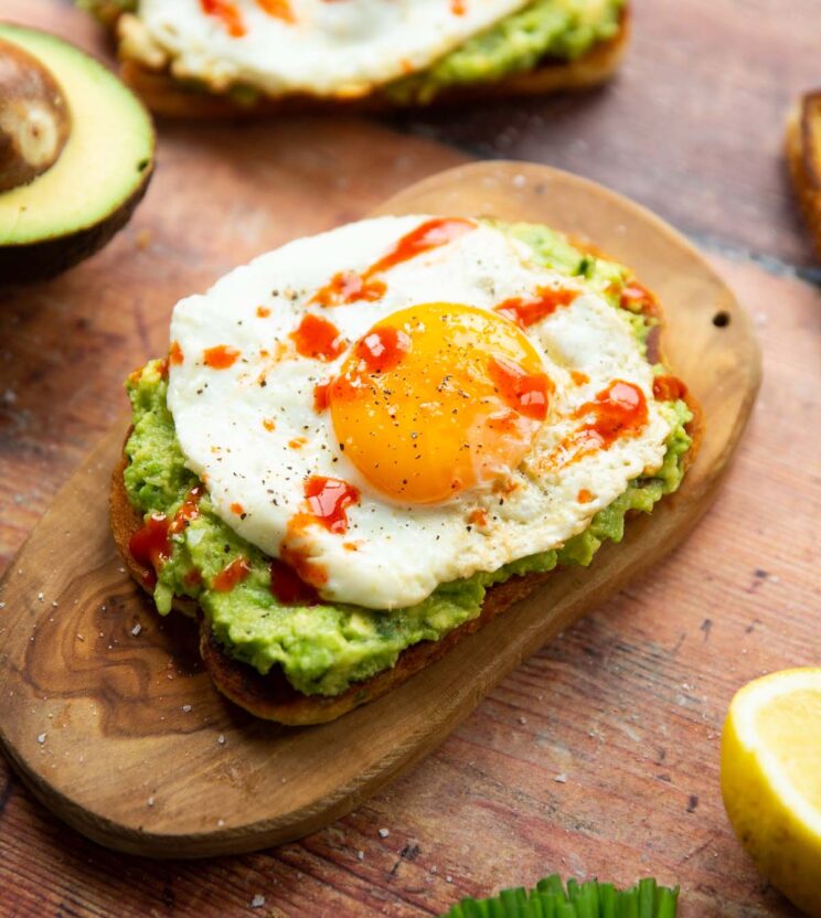 close up shot of avocado egg toast on small chopping board surrounded by garnish