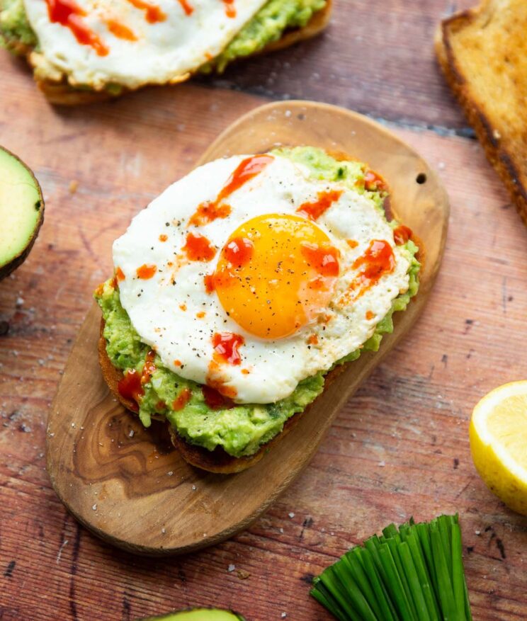 avocado fried egg toast on small wooden chopping board surrounded by garnish on wooden surface