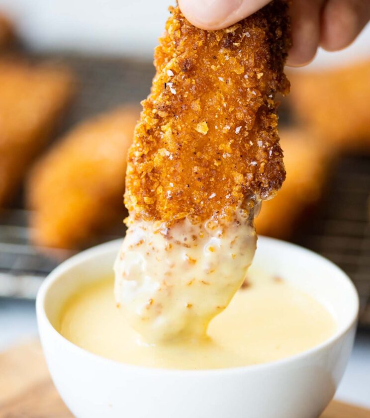 hand dunking a cornflake crusted chicken tender into small white pot of honey mustard dip