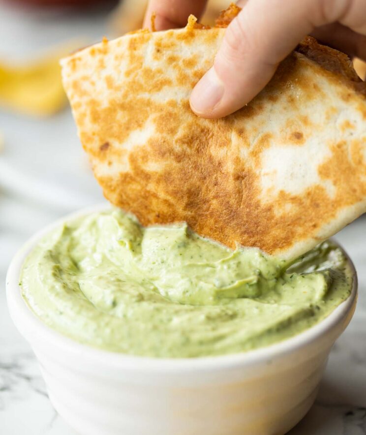 hand dunking half a refried bean quesadilla into small pot of avocado dip