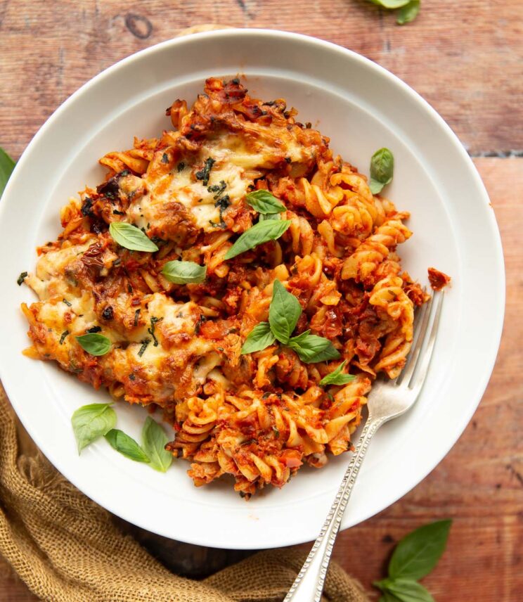 overhead shot of tomato tuna pasta bake in white bowl with silver fork garnished with basil on wooden surface