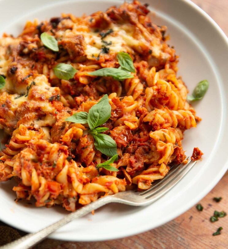close up side shot of tomato tuna pasta bake in white bowl with silver fork garnished with basil
