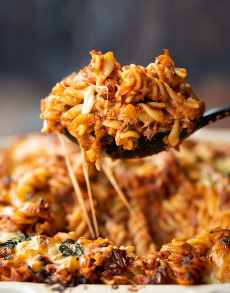 close up shot of black serving spoon scooping up tuna pasta bake out of white baking dish