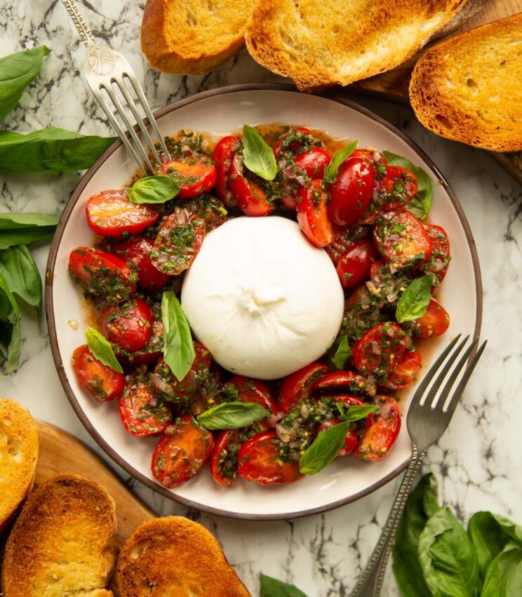 overhead shot of burrata tomato salad surrounded by fresh basil, toast and 2 silver forks