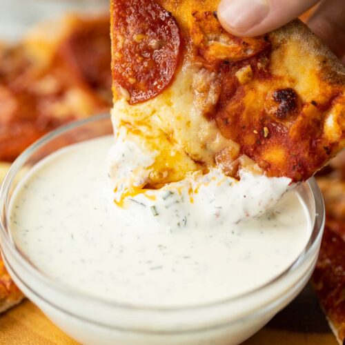 close up shot of hand dunking pepperoni pizza into glass bowl of ranch