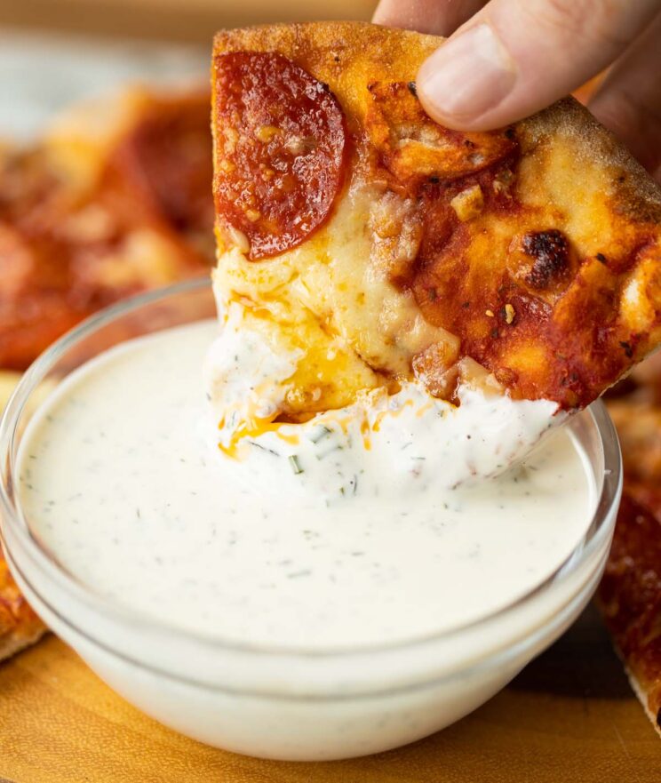 close up shot of hand dunking pepperoni pizza into glass bowl of ranch