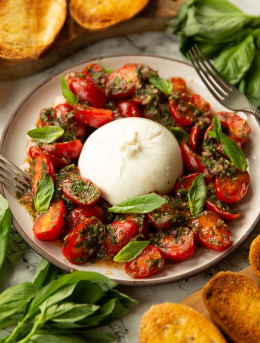 tomato burrata salad served on small white plate surrounded by fresh basil, toasted bread and 2 silver forks