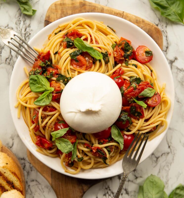 over shot of caprese burrata pasta on white plate with chopping board and two silver forks