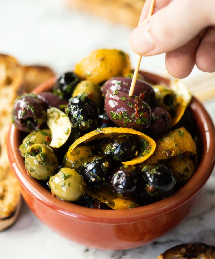 close up shot of marinated olives in small brown dish with hand poking toothpick into one