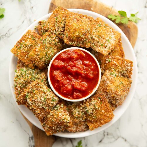 overhead shot of deep fried ravioli on small white plate surrounding a pot of marinara dip