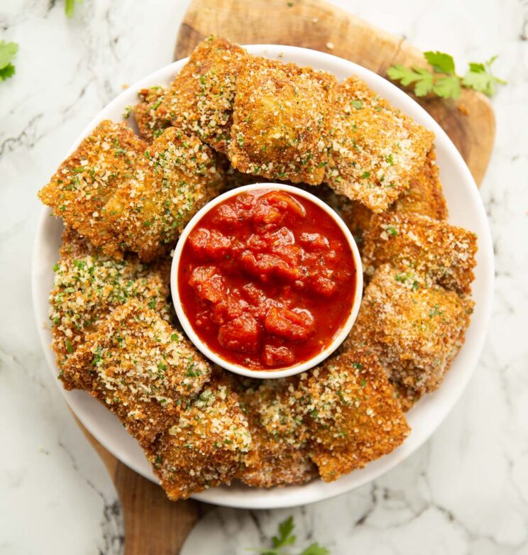 overhead shot of deep fried ravioli on small white plate surrounding a pot of marinara dip