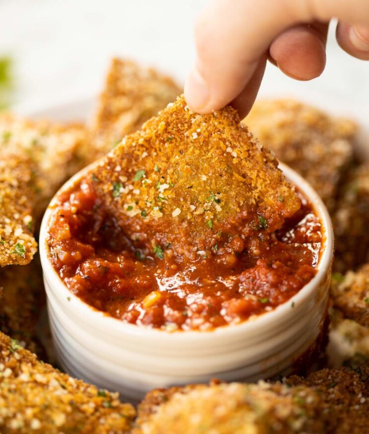 hand dunking one piece of fried ravioli into small white pot of marinara dip