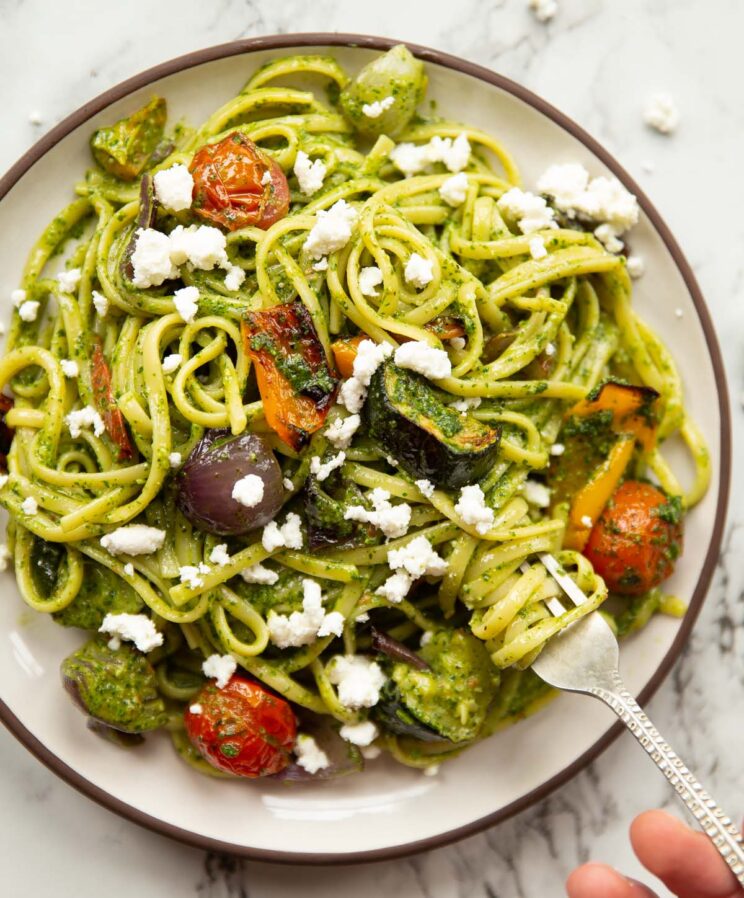 overhead shot of roasted vegetable pesto pasta on small white plate with hand twisting in silver fork