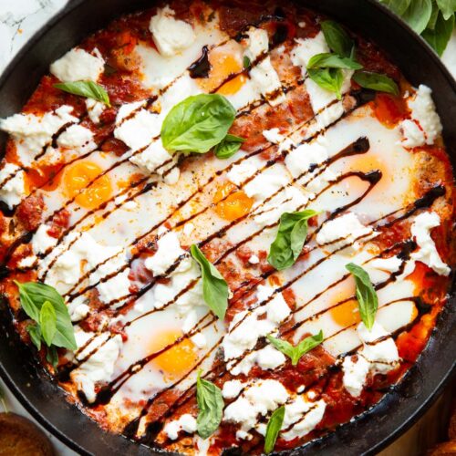 overhead shot of caprese baked eggs in skillet surrounded by toast and fresh basil