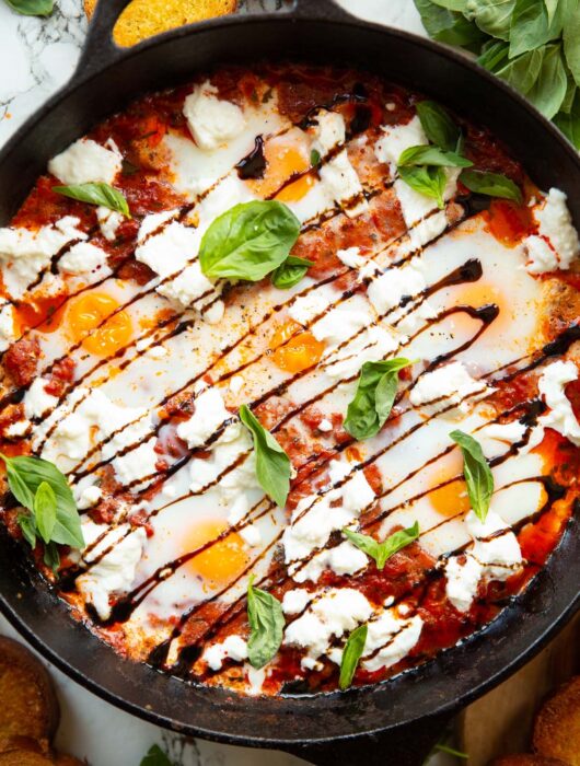 overhead shot of caprese baked eggs in skillet surrounded by toast and fresh basil
