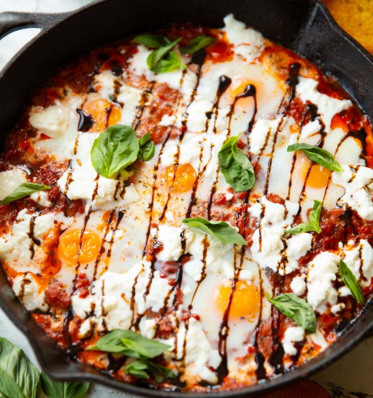 close up shot of caprese eggs in skillet surrounded by toast and fresh basil