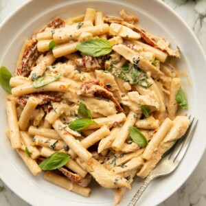 overhead shot of chicken sun dried tomato pasta in white dish with silver fork garnished with fresh basil