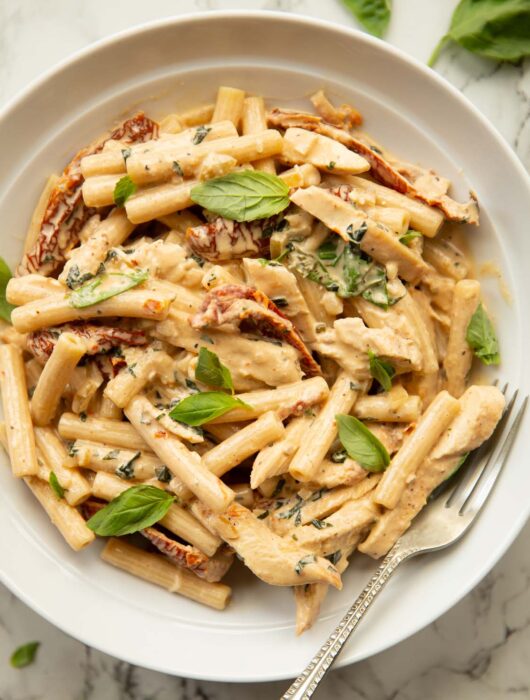 overhead shot of chicken sun dried tomato pasta in white dish with silver fork garnished with fresh basil